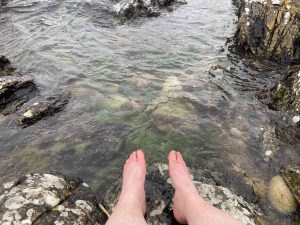 A Scottish rock pool with clear grean tinted water, bare feet in foreground. They are slightly reddened, indicating the water is cold